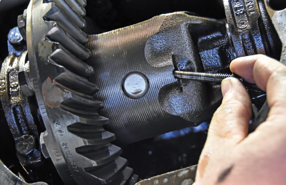 Extreme close-up of a differential gear assembly. A hand is removing a small threaded pin shaft bolt from the center section near the ring gear.
