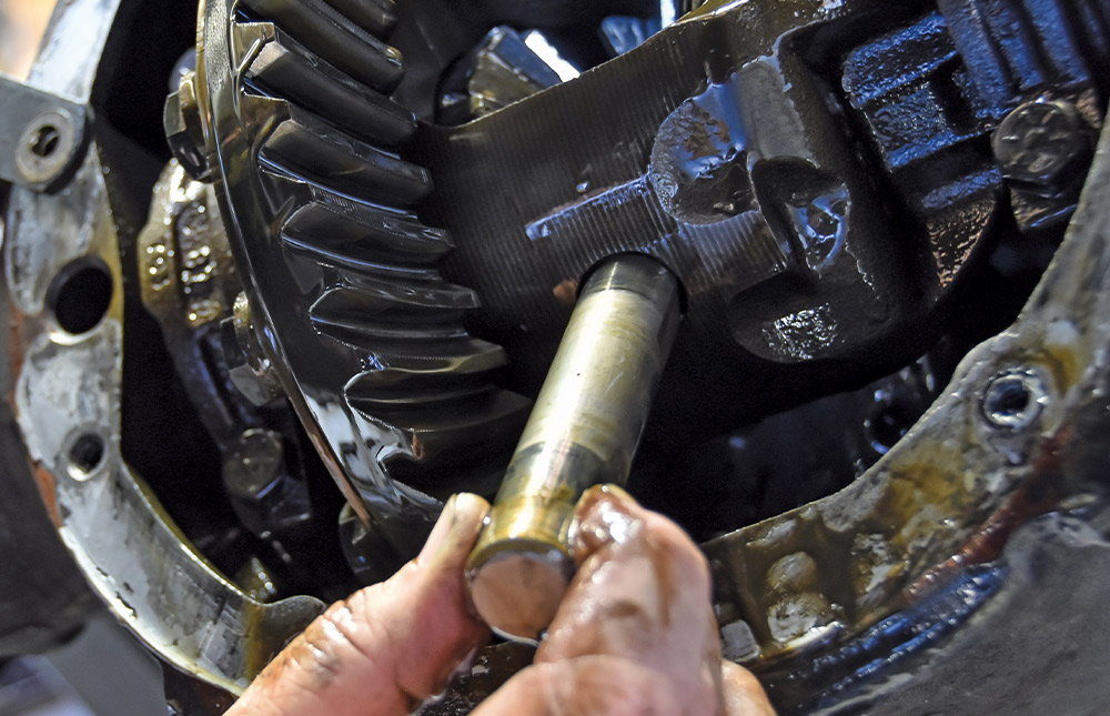 A hand removes a cylindrical cross-pin shaft from the center section of an oily differential gear assembly.