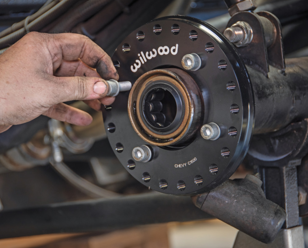 A hand inserts a bolt into a black circular Wilwood adapter plate mounted on an axle housing. The plate is labeled "CHEVY C1500".