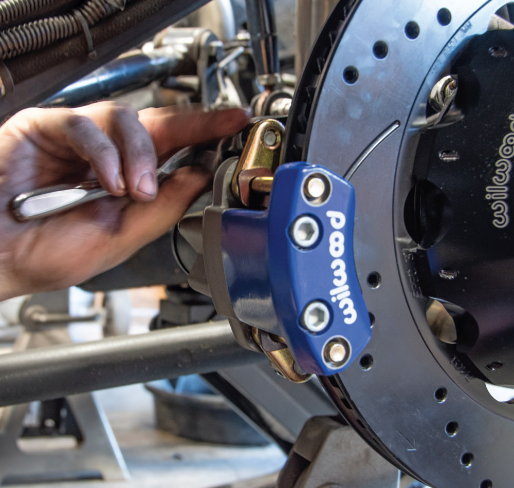 A hand uses a wrench to adjust or tighten the mounting hardware for a small blue Wilwood electronic parking brake caliper. The caliper is mounted to the side of a large black slotted and drilled disc brake rotor.