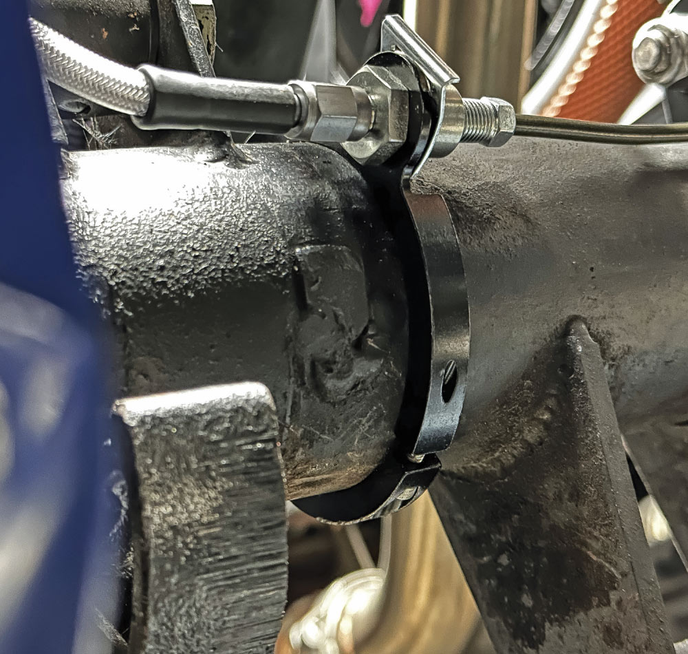 Close-up of a black Wilwood aluminum clamp-on tab secured around a vehicle axle, holding a brake line fitting.