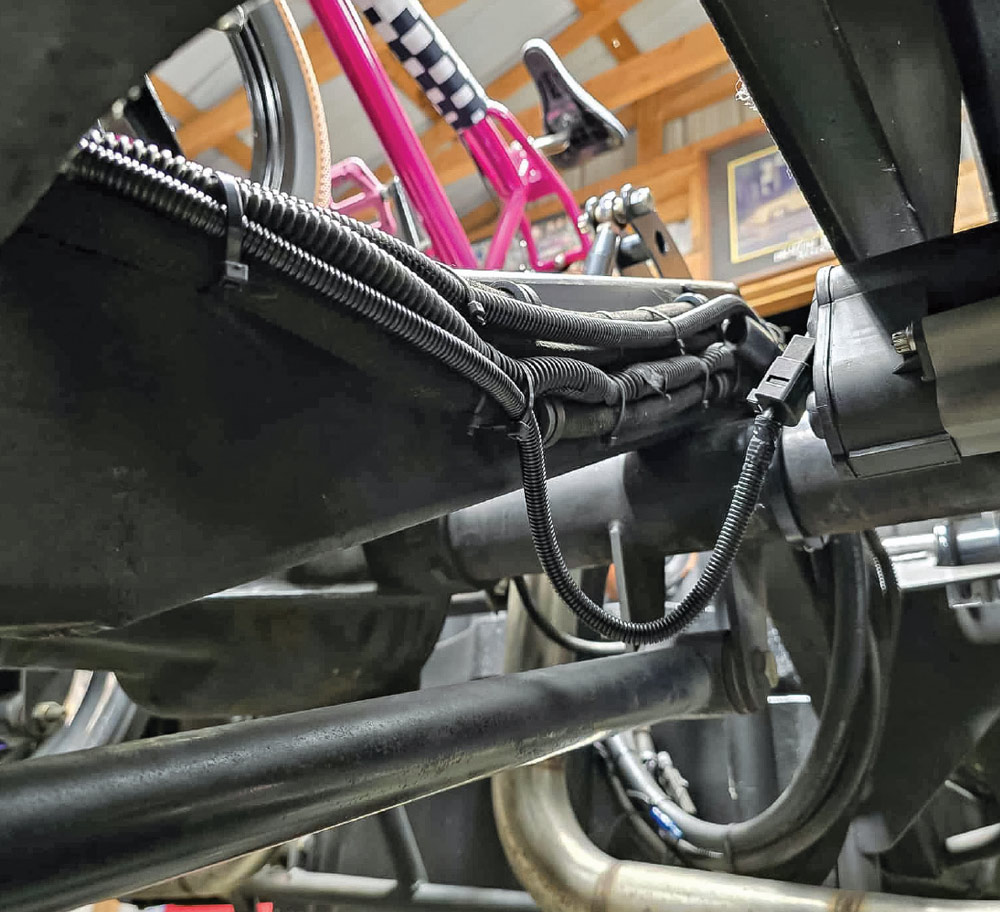 Black corrugated wiring looms are zip-tied along a metal frame rail on the underside of a vehicle. A section of the loom loops down toward the suspension components.
