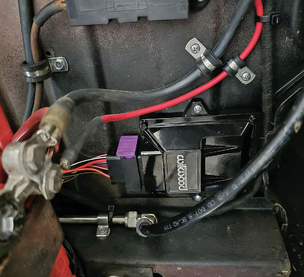 A black Wilwood control module mounted to a vehicle panel. A wiring harness with a purple connector is plugged in. Red and black wires are routed nearby with clamps.
