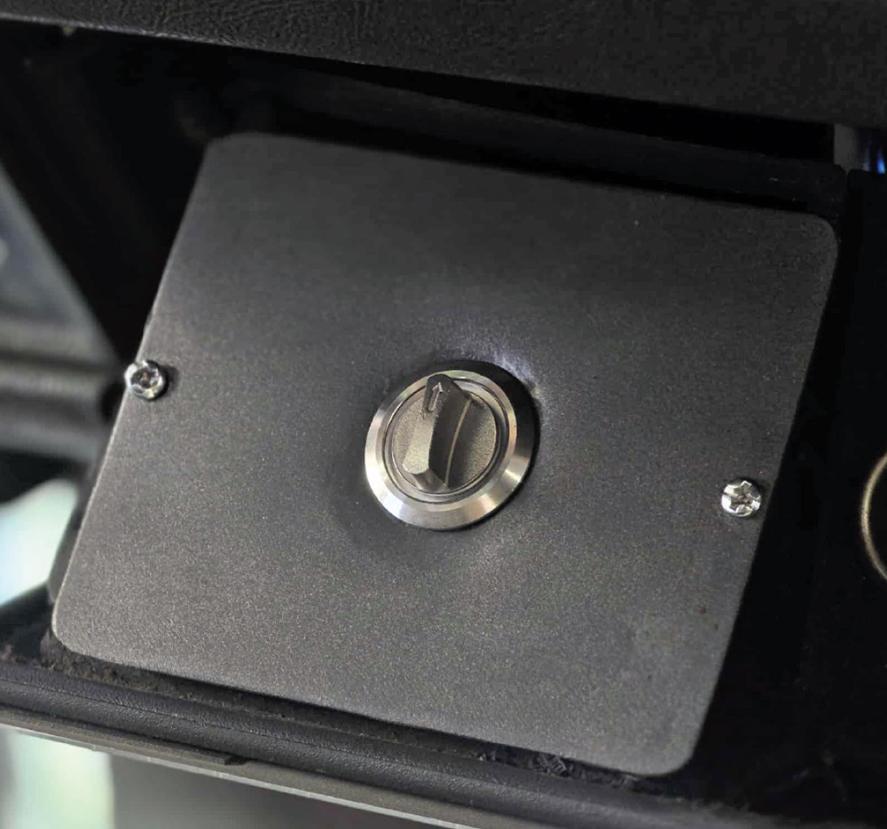 A circular metal switch with a small toggle is mounted on a black panel secured by two screws.