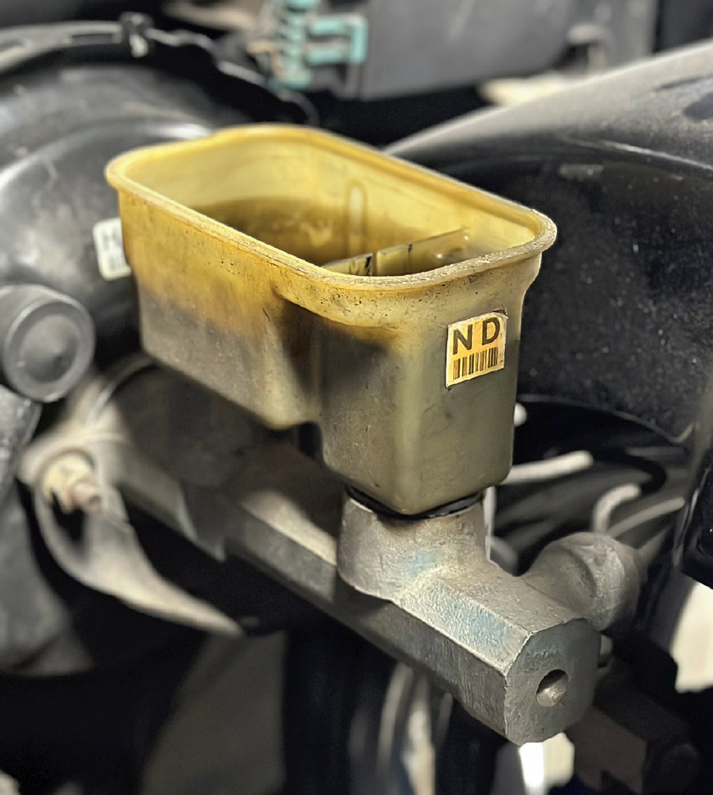A weathered translucent plastic brake fluid reservoir with an "ND" label is mounted on a metal master cylinder.