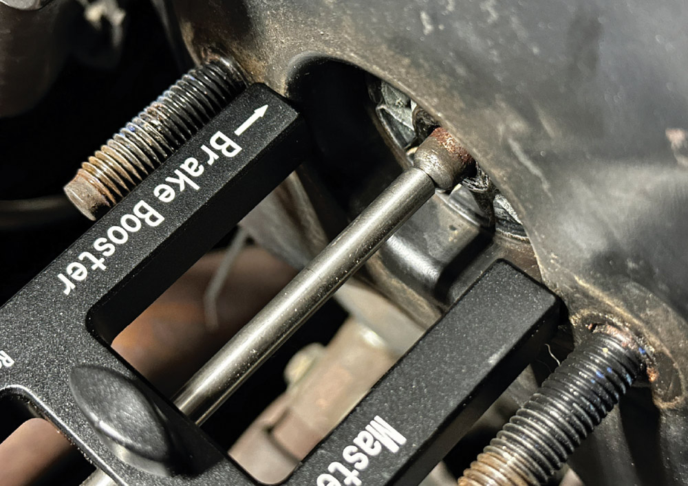 A black Wilwood depth measurement tool is placed against a brake booster to measure the push rod. The tool has "Brake Booster" and an arrow printed on it.