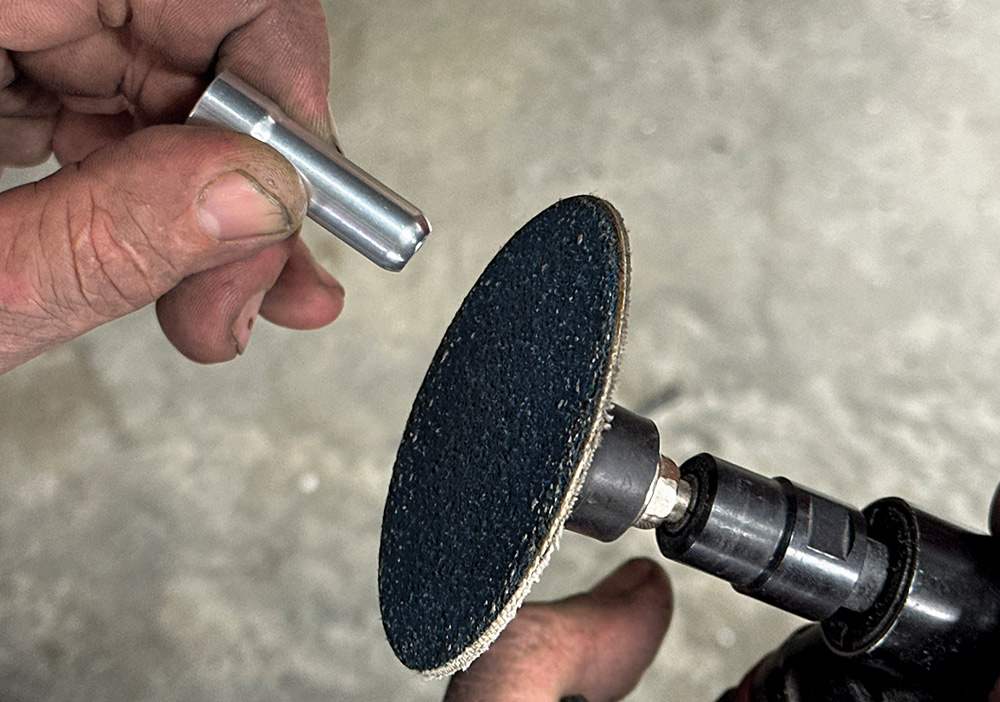 A hand holds a small silver metal pin near the surface of a black sanding disc on a handheld power tool.