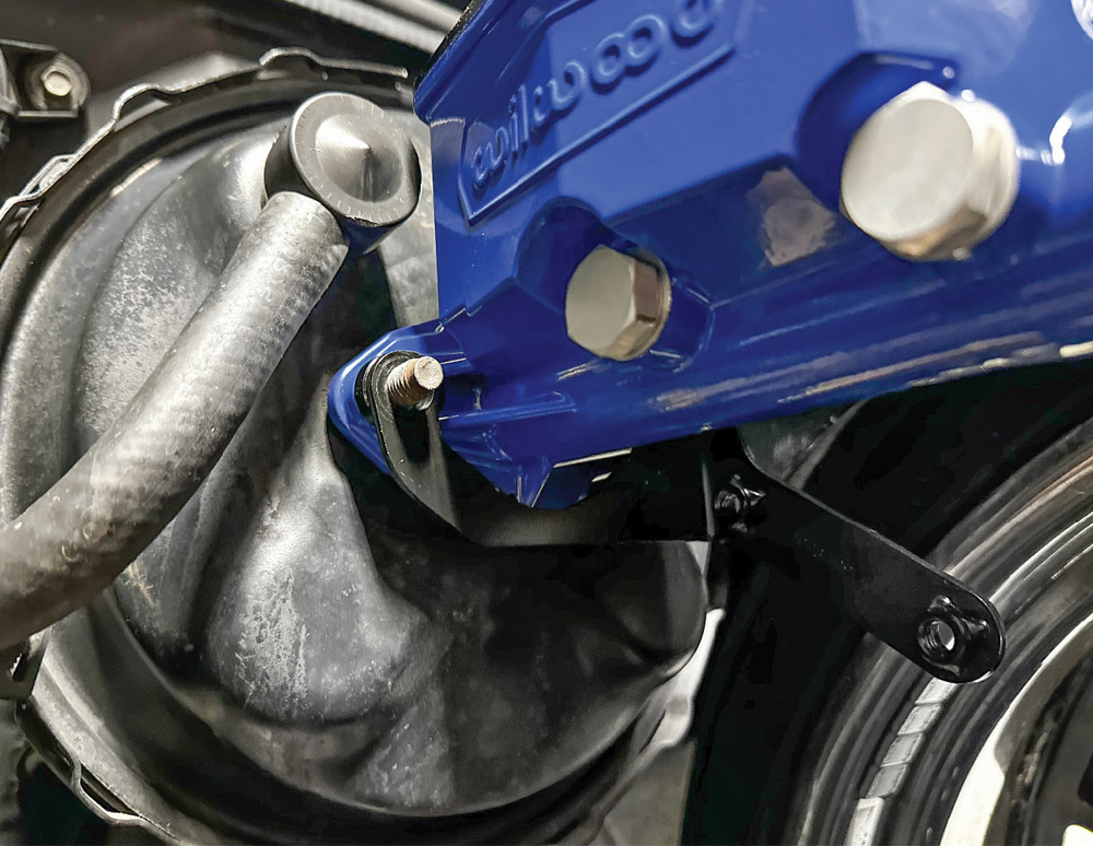 A blue Wilwood master cylinder is mounted onto a black brake booster with a black metal proportioning valve bracket in between.