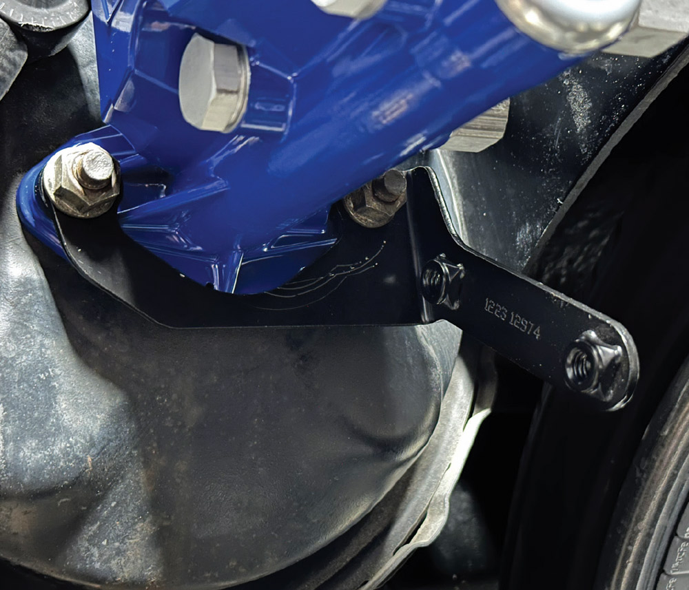 Close-up of the base of a blue Wilwood master cylinder secured to a black booster with nuts and washers. A black metal bracket is held by the same hardware.