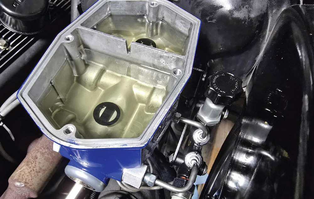 Top-down view of an open blue master cylinder reservoir with two fluid-filled compartments. Silver mounting nuts and washers are on the booster studs below. A silver brake line is attached.