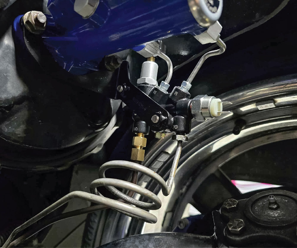 A black proportioning valve is mounted below a blue master cylinder. Multiple silver brake lines are connected to it, including one with a coiled loop. A brass reducer fitting is visible.