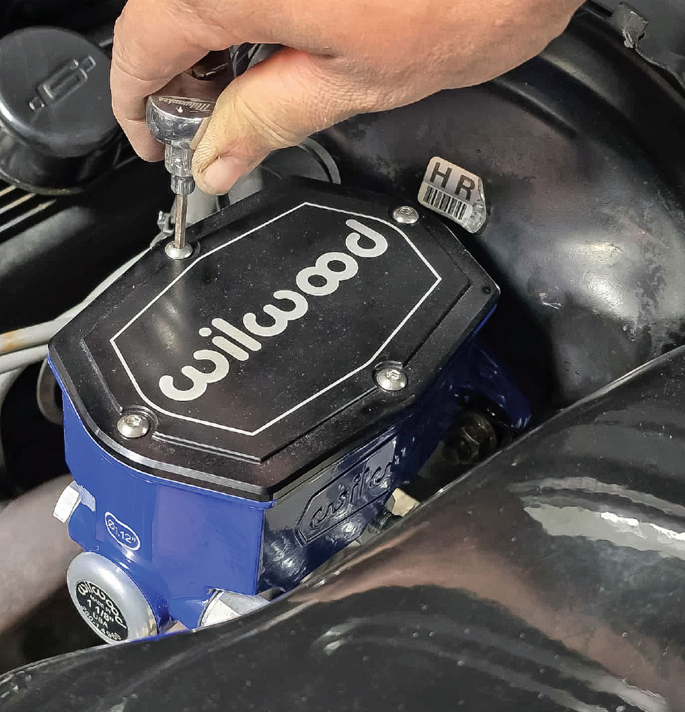 A hand uses a small tool to tighten a screw on the black lid of a blue Wilwood master cylinder.