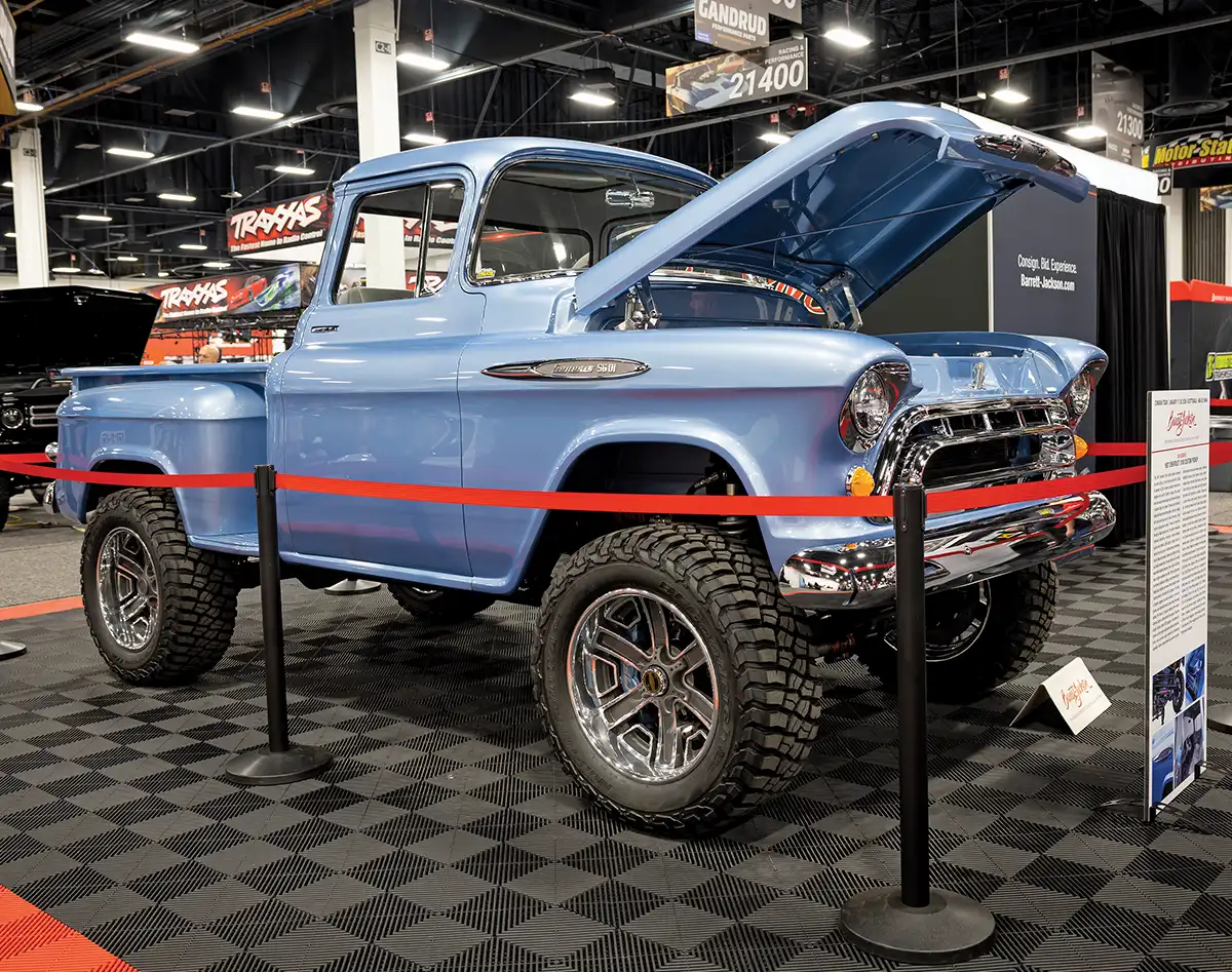 A side profile of a restored 1957 Chevrolet 3100 "Task Force" pickup truck on display at a show, featuring a metallic light blue paint job, lifted 4x4 suspension, and chrome off-road wheels