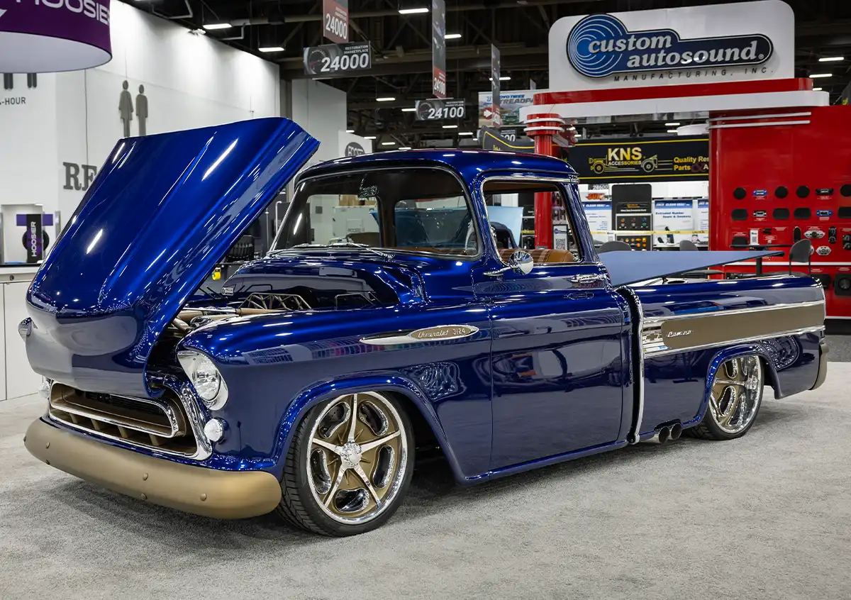 A front three-quarter view of a custom 1957 Chevrolet 3100 Cameo pickup truck on display at SEMA, featuring a deep metallic blue paint job, slammed suspension, gold-finished bumpers, and modern five-spoke wheels