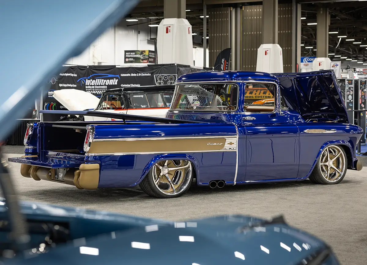 A rear three-quarter view of a custom 1957 Chevrolet 3100 Cameo restomod on display at SEMA, featuring a deep metallic blue finish, gold-painted bumpers, a slammed suspension, and integrated side-exit exhaust