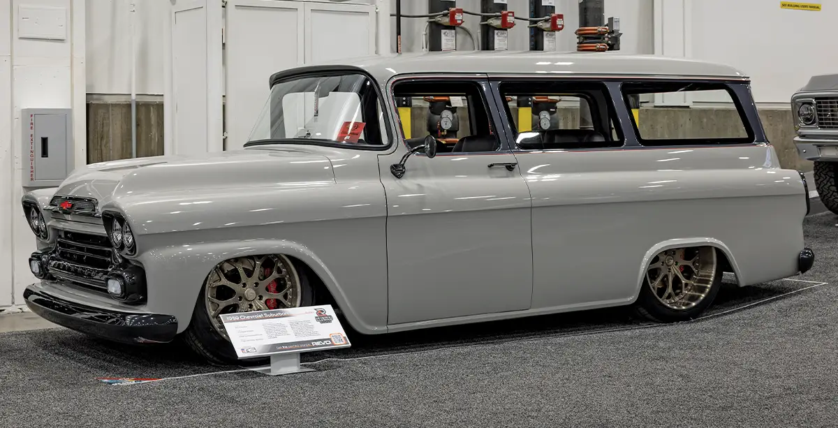 A side profile of a custom 1959 Chevrolet Suburban restomod on display at an indoor car show, featuring a glossy grey paint job, slammed suspension, and large bronze multi-spoke wheels