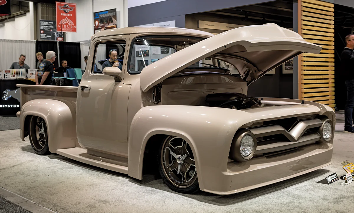 A front three-quarter view of a custom 1953-1956 Ford F-100 pickup truck on display at a car show, featuring a monochrome tan paint job, slammed suspension, and modern five-spoke wheels with a matching tan finish