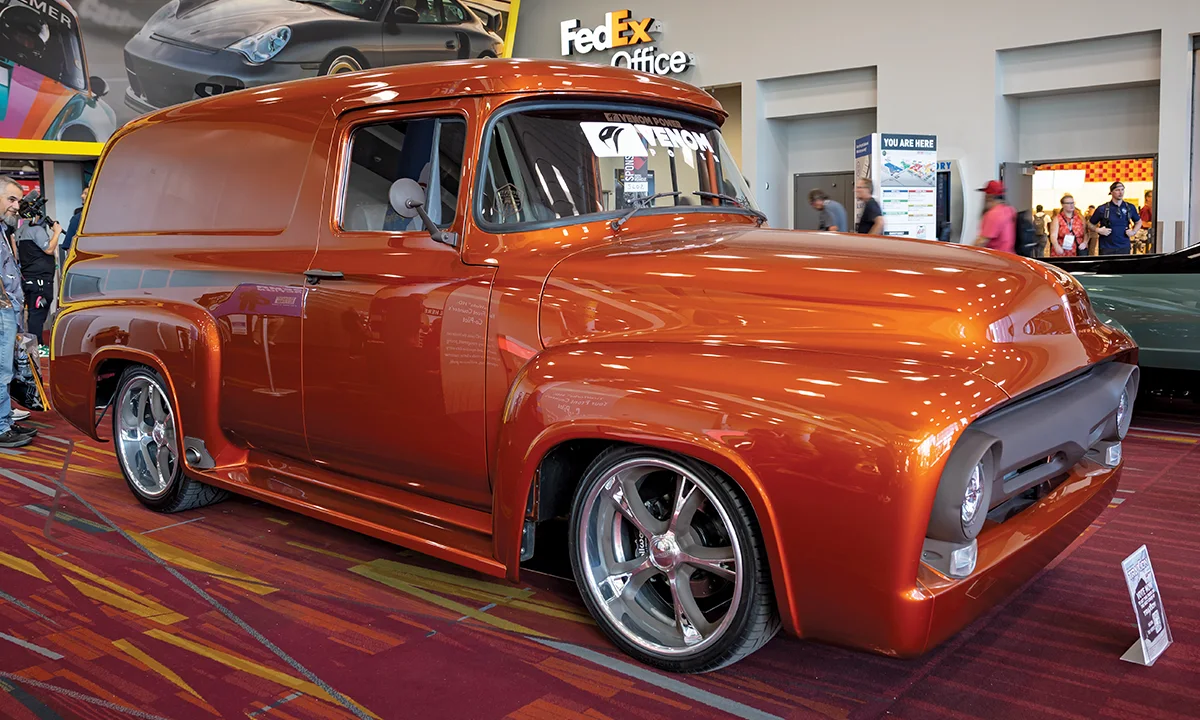 A front three-quarter view of a custom metallic orange 1953-1956 Ford F-100 panel delivery van on display at SEMA 2025, featuring a slammed suspension, modern five-spoke chrome wheels, and a matte gray grille