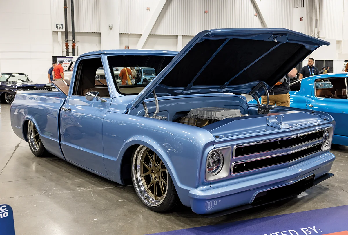 A front three-quarter view of a custom metallic light blue Chevrolet C10 pickup truck on display at a car show, featuring an ultra-low slammed suspension, deep-dish bronze multi-spoke wheels, and a performance engine with the hood open