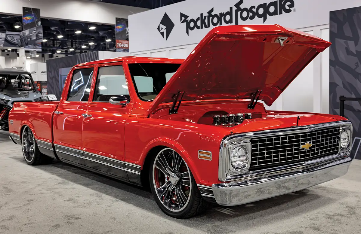 A front three-quarter view of a custom metallic red Chevrolet C10 crew cab pickup truck on display at SEMA, featuring a slammed suspension, large chrome and red multi-spoke wheels, and a performance engine with the hood open