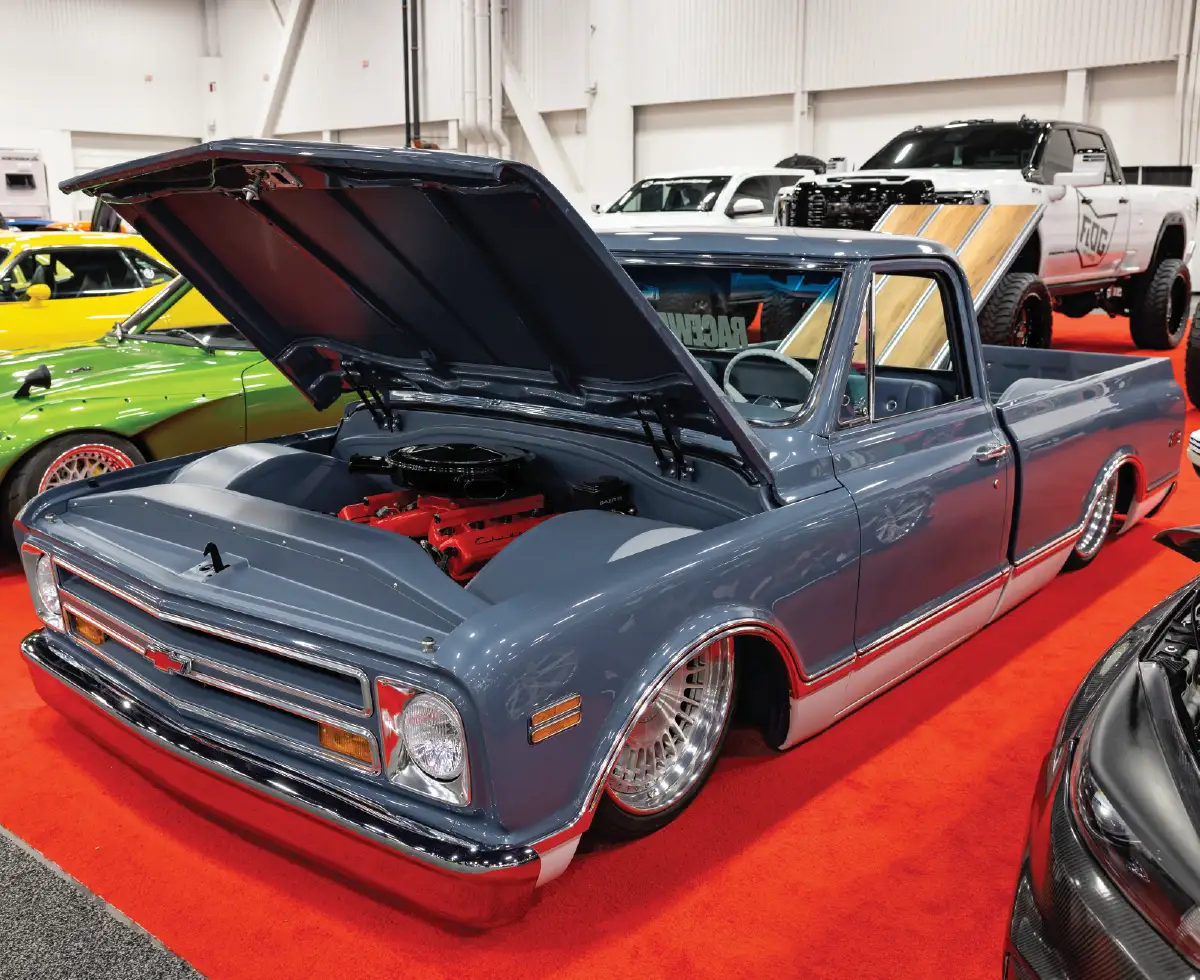 A front three-quarter view of a custom slate grey 1967-1972 Chevrolet C10 pickup truck on display at a car show, featuring an ultra-low slammed suspension, a custom red-block engine with the hood open, and a wood-lined truck bed tilted upward
