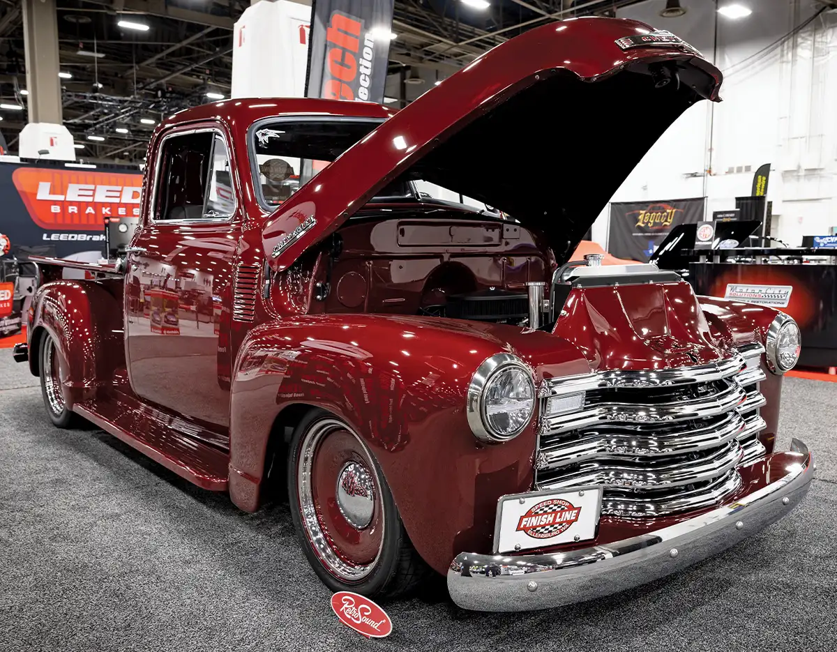 A front three-quarter view of a restored 1947-1954 Chevrolet Advance Design pickup truck on display at a car show, featuring a deep maroon paint job, slammed suspension, and chrome five-bar grille with the hood open