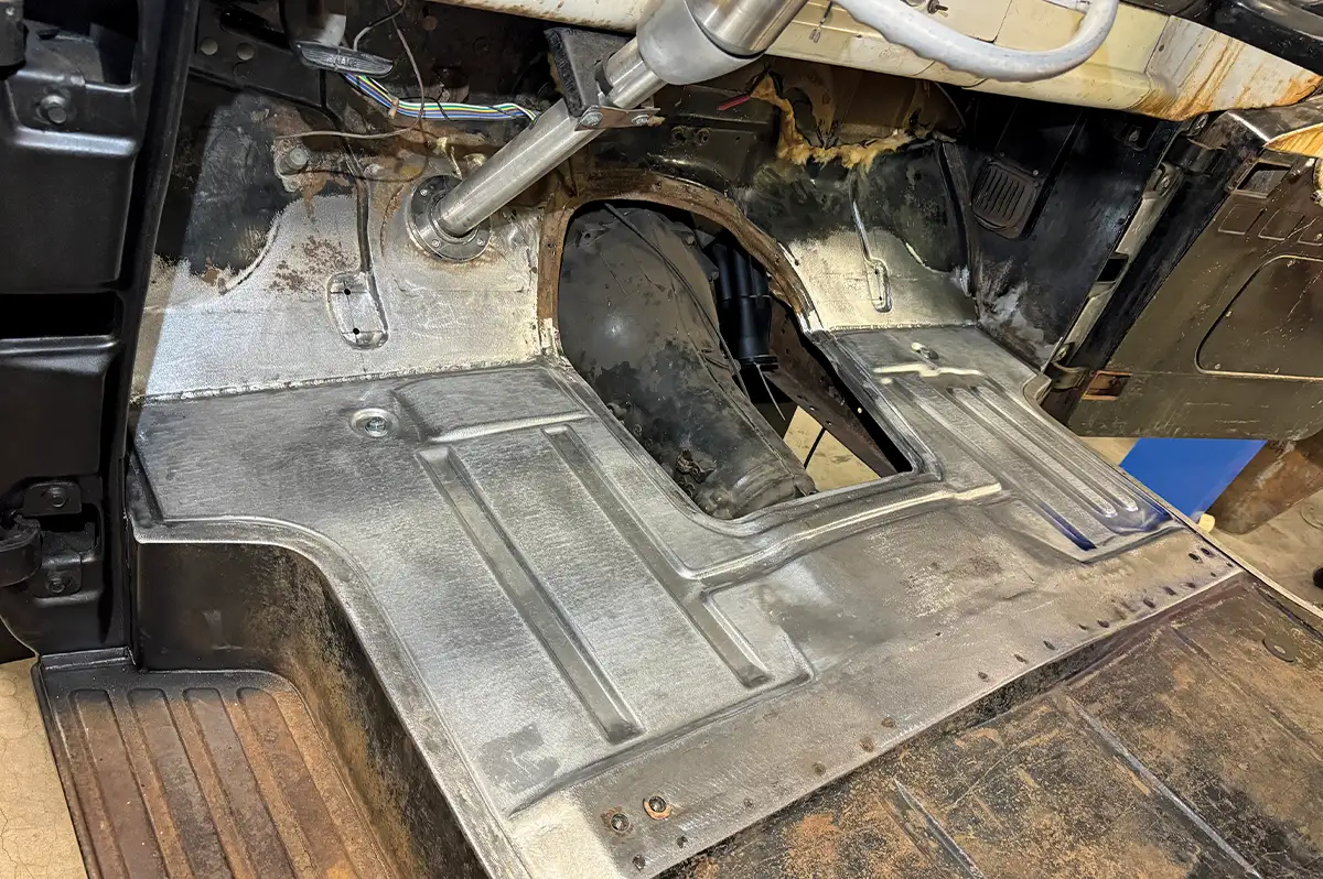 A high-angle interior shot of a classic truck restoration project, featuring newly fabricated and welded raw steel floor pans and a raised transmission tunnel to accommodate a modern drivetrain upgrade