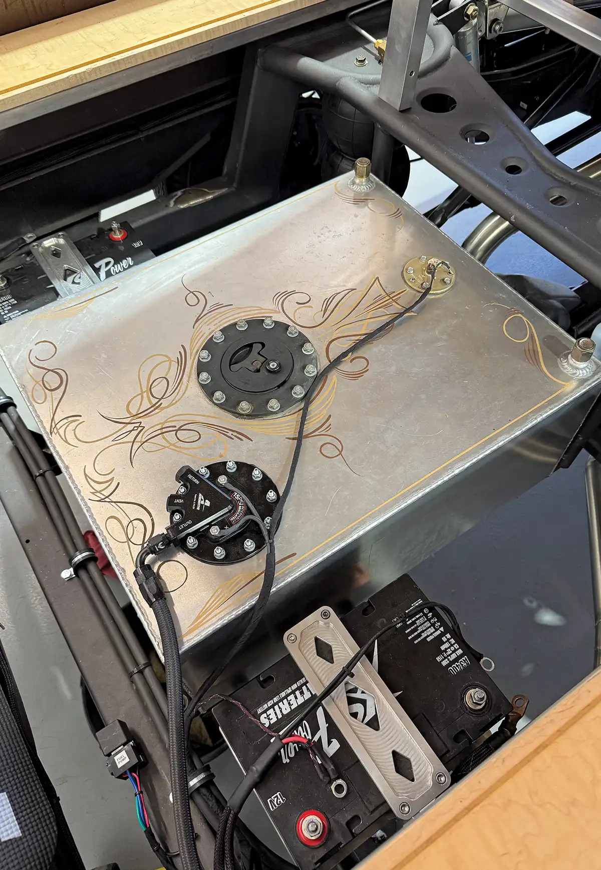 A top-down view of a custom brushed aluminum fuel cell with intricate gold pinstriping, an Aeromotive fuel pump, and a high-performance XS Power 12V battery mounted within a fabricated vehicle chassis