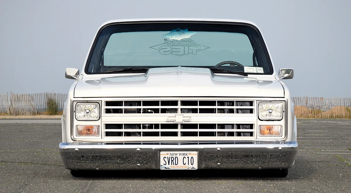 A head-on front view of a custom white 1980s Chevrolet C10 "Squarebody" pickup truck parked in an open lot, featuring a slammed suspension, chrome bumper, and a custom "SVRD C10" New York license plate