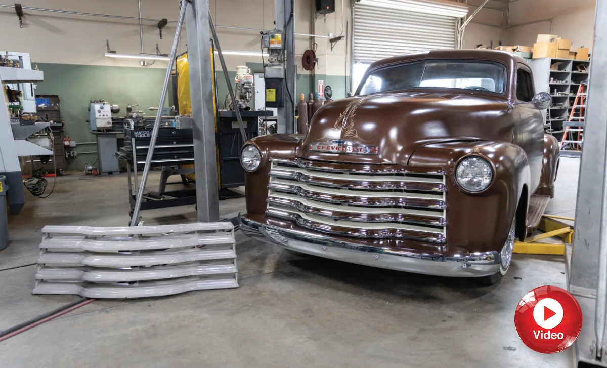 A professional shop photograph of a show-quality Chevy 3100 restomod with a satin brown finish, showcasing a lowered ride height, white-wall tires, and an extra painted grille sitting on the floor nearby