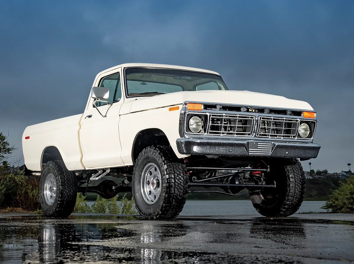 A front three-quarter view of a lifted white vintage 1970s Ford F-150 pickup truck parked on wet pavement, featuring a chrome front bumper with an American flag emblem, off-road tires, and a white-out grille
