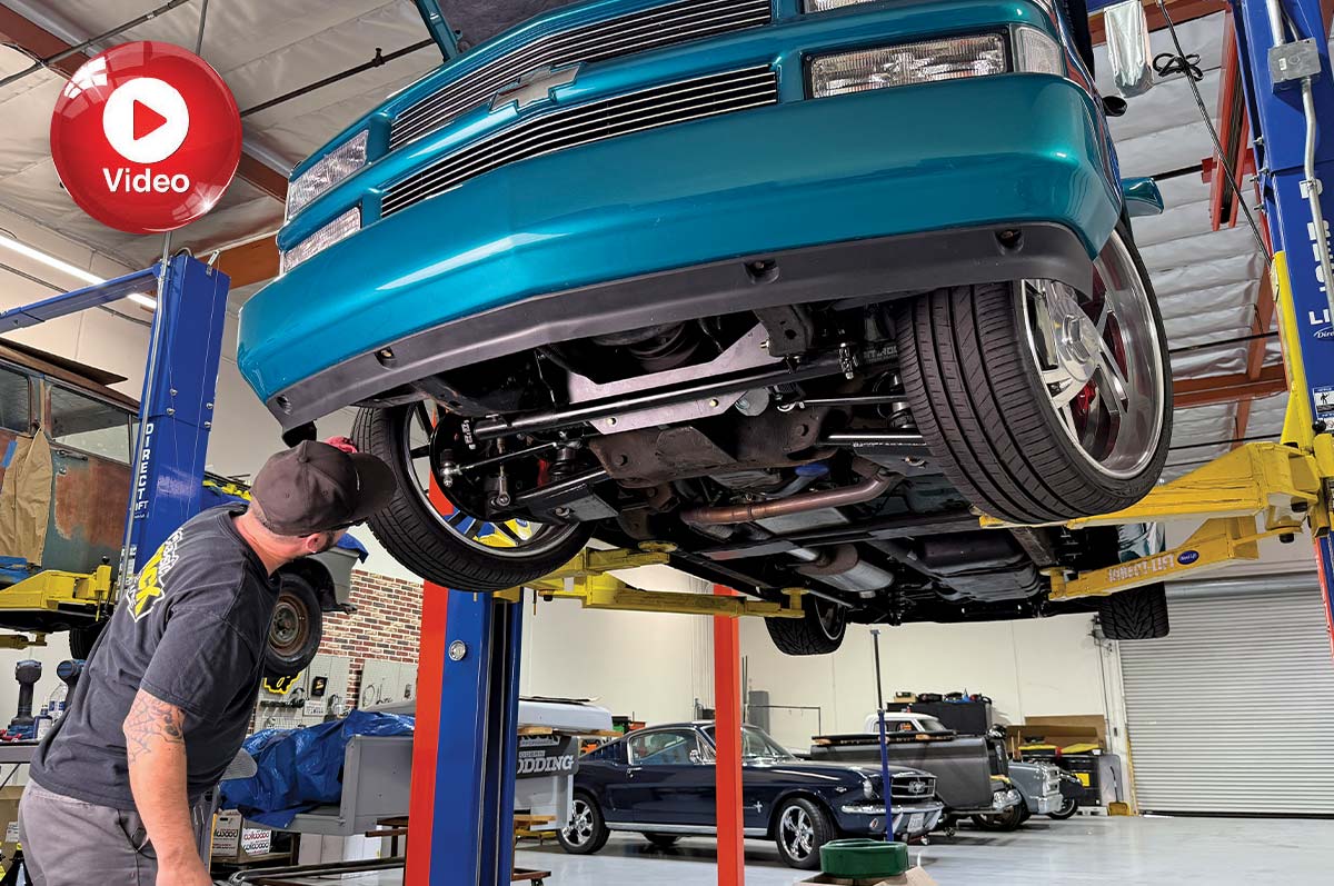 A mechanic looks up at the undercarriage of a teal truck raised on a hydraulic lift inside an auto shop. Classic cars are parked in the background, and a red "Video" play button overlays the top left corner.
