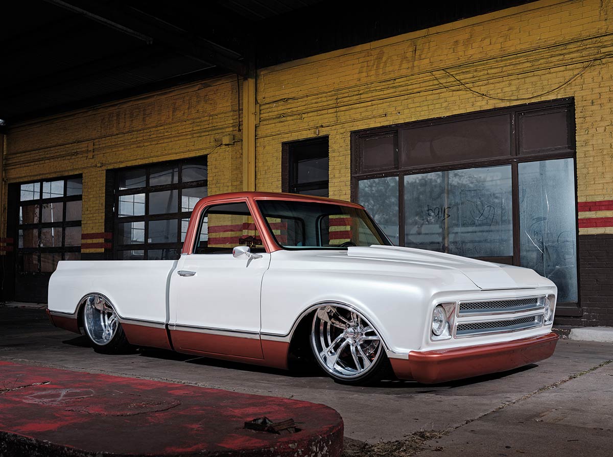 A custom, extremely lowered white pickup truck with copper accents on the roof and lower trim is parked outside a weathered yellow brick building. The truck features large polished chrome wheels.
