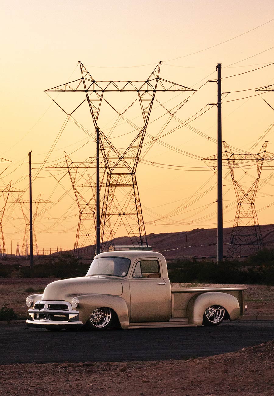A champagne-gold custom vintage pickup truck lowered to the ground, parked near power line towers against a desert sunset.