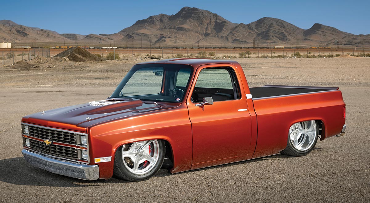 A custom metallic copper-orange lowered pickup truck parked outdoors with desert mountains in the background under a blue sky.