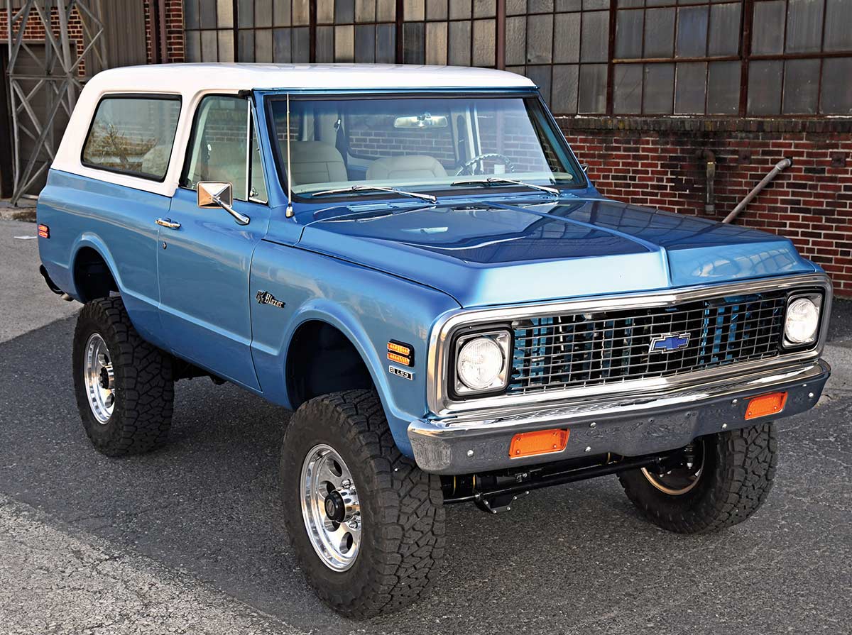A restored classic blue Chevrolet K5 Blazer with a white hardtop roof and off-road tires parked by a brick building.