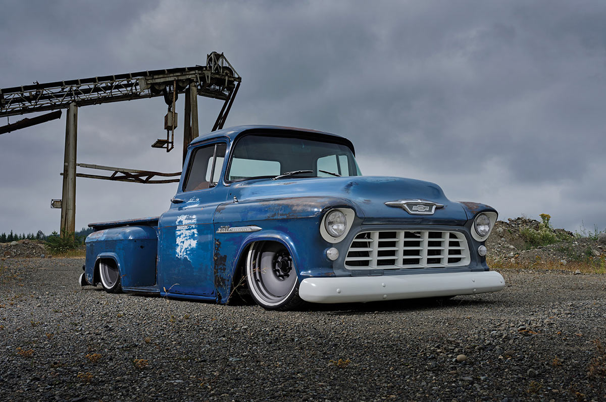 A custom lowered blue vintage Chevrolet pickup truck with white wheels parked on a gravel lot in front of industrial machinery.